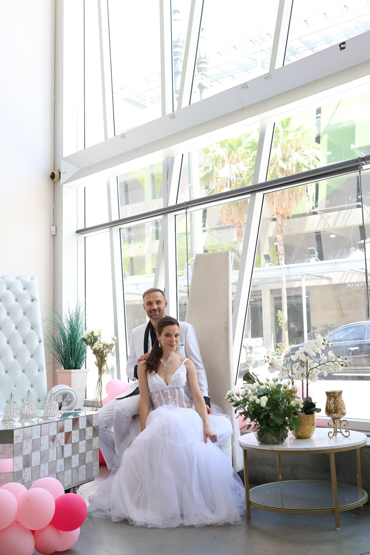 A couple in wedding attire seated in the sunlit glass atrium